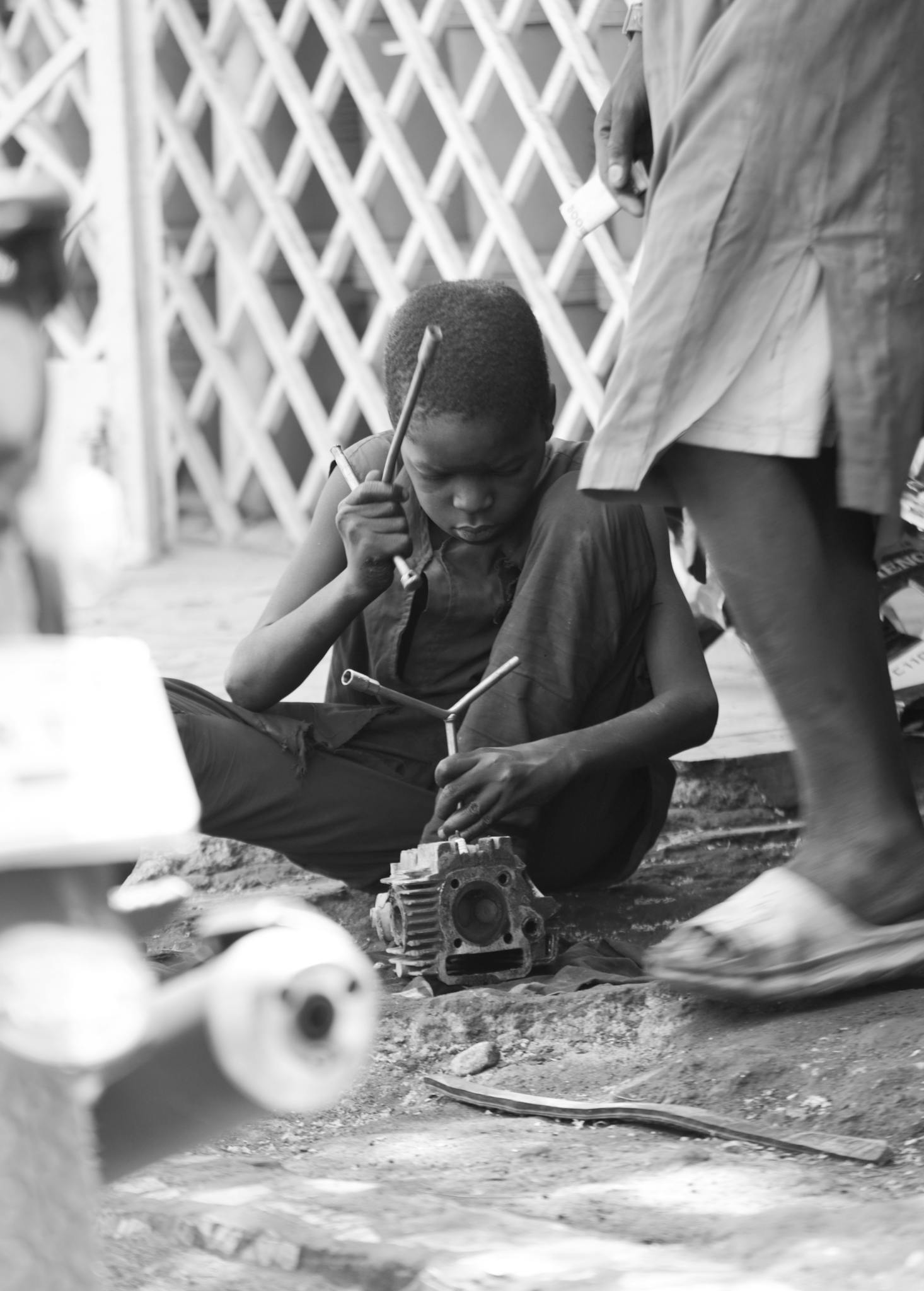Black and white image of a young boy focused on repairing equipment outdoors.